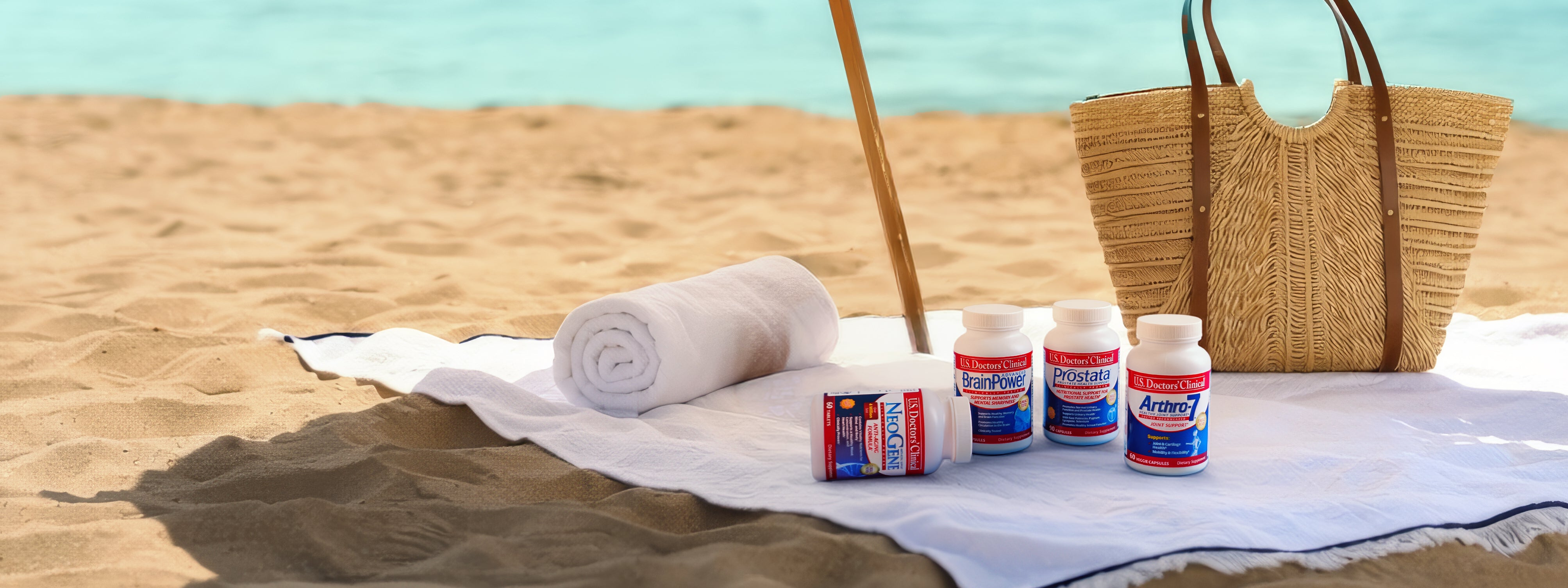Four dietary supplement bottles branded "U.S. Doctors' Clinical" sit on a white towel spread across a sandy beach. A rolled-up white towel rests nearby, and a large woven wicker tote bag with leather handles stands in the background. The calm, turquoise ocean water is visible under the bright sunlight.